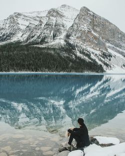 Man sitting at lake by snowcapped mountains during winter