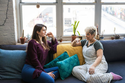 Female roommates talking while relaxing together on sofa against window at home