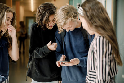 Smiling male students using smart phone while standing by female friends in school corridor