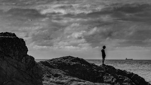 Silhouette boy standing on rock by sea against sky