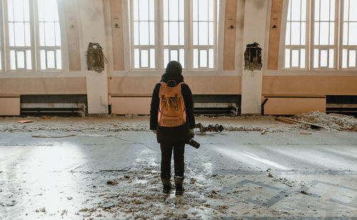 Rear view of young woman with backpack and camera standing in abandoned room