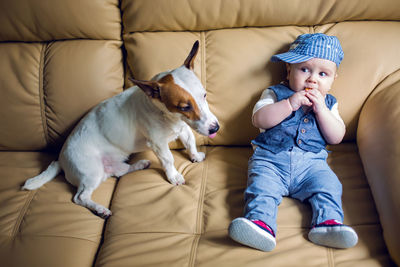 Gay boy kid blonde in a cap , pants, tie and vest sits with jack russell terrier dog on sofa