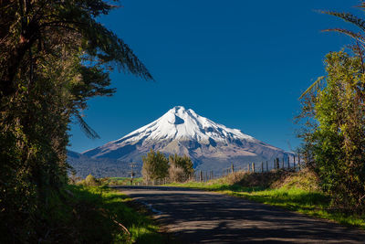 Empty road leading towards snowcapped mountain against clear blue sky