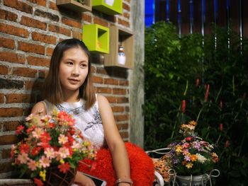 Woman looking away while sitting with flowers against brick wall