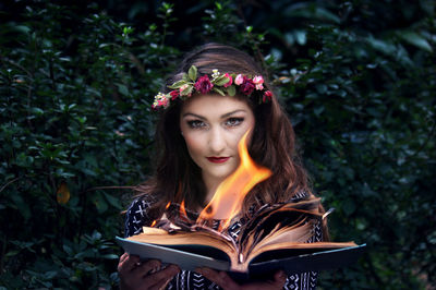 Portrait of young woman holding burning book standing against plants