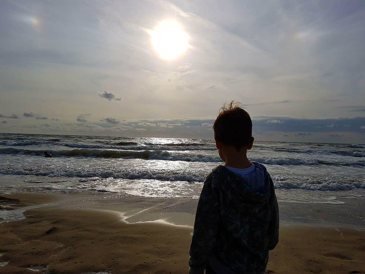 REAR VIEW OF BOY STANDING AT BEACH DURING SUNSET