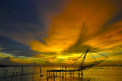 Silhouette chinese fishing net in sea against dramatic sky during sunset