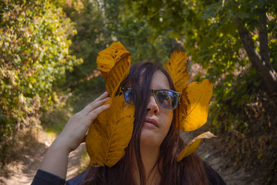 Portrait of young woman holding sunglasses against trees