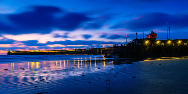 Illuminated building by sea against sky at dusk