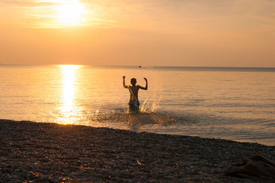 Young boy playing on shore against sky during sunset