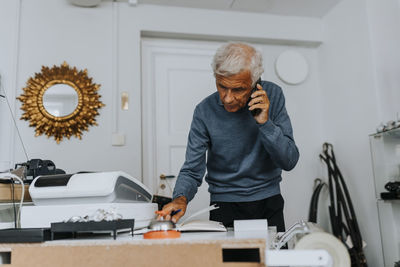 Senior male entrepreneur writing in diary while talking on smart phone in antique shop