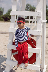 Child boy in striped clothes and red pants  standing on beach. white lifeguard tower, with a circle