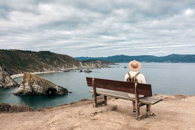 Back view high angle of male explorer in hat sitting on wooden bench and admiring spectacular scenery of sea and rocks