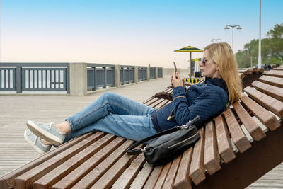 Young lonely blonde woman on empty city embankment in sun lounger uses smartphone