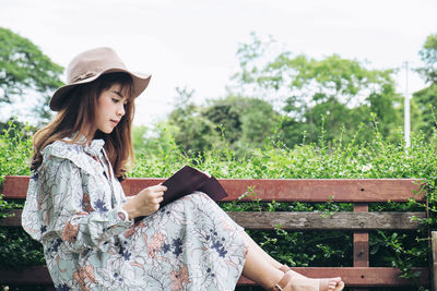 Woman reading book while sitting on bench by plants