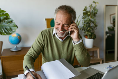 Smiling man writing in book while talking on mobile phone at home