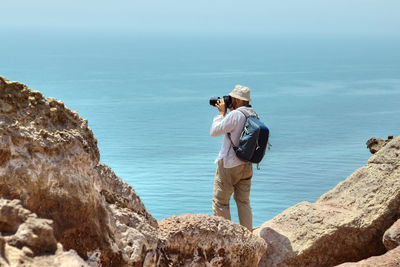 Man photographing with camera while standing on rock formation against sea