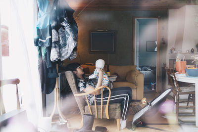 Mother playing with toddler while sitting on chair seen though window