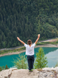 High angle view of boy standing with arms raised on stone against lake