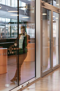 Full length of businesswoman talking on smart phone seen through glass door in office