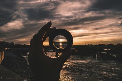 Close-up of silhouette hand holding camera lens against sky