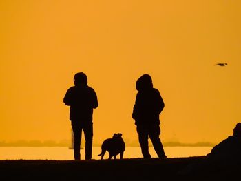 Silhouette friends standing with dog at lakeshore against clear orange sky