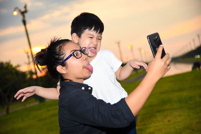 Playful mother and son sticking out tongues while taking selfie with smart phone on field during sunset