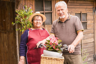 Portrait of senior couple standing with potted plant in bicycle basket against house