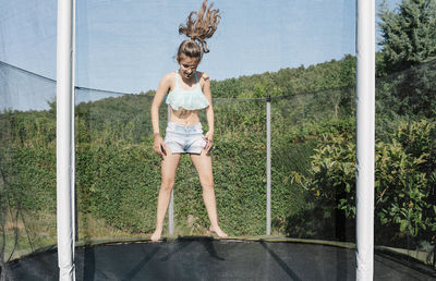 Horizontal photo of a smiling young brunette teenager jumping on a trampoline with net around on the green yard outdoors. the girl wears short jeans and a top and looks fun, active