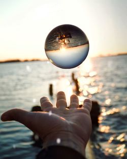 Cropped hand catching crystal ball against sea during sunset