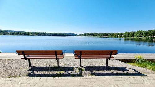 Empty bench by lake against clear blue sky