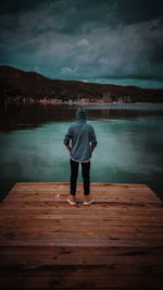Rear view of teenage boy standing on pier over lake against sky