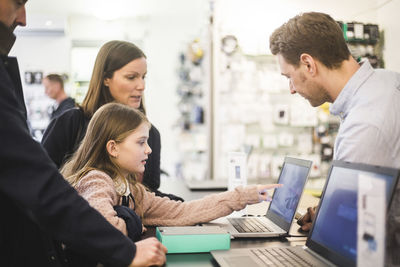 Cute girl pointing at laptop's screen to salesman while standing with family in computer store