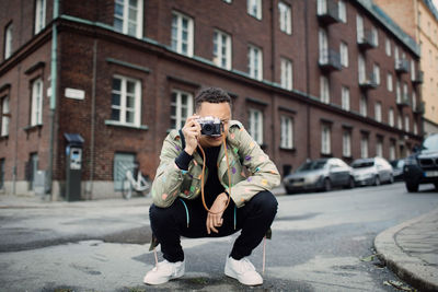 Young male photographer crouching while photographing on street against building in city