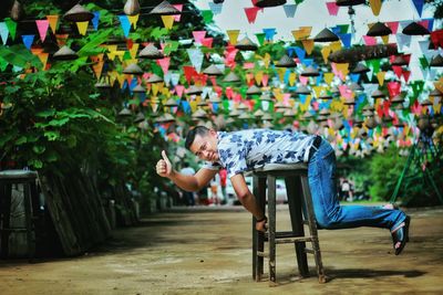 Full length portrait of man gesturing while leaning on stool against prayer flags