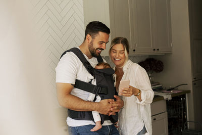 Smiling woman sharing smart phone with man carrying son in kitchen at home