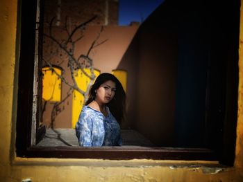 Portrait of young woman looking away while sitting on window