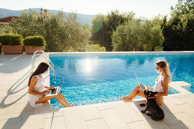 High angle view of women using laptop while sitting by swimming pool with dog