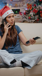 Young woman talking on phone while sitting on sofa watching tv at home