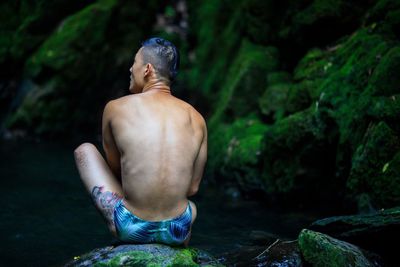 Rear view of shirtless man sitting on rock in forest