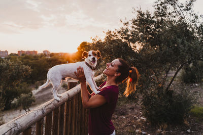 Smiling woman with dog by railing during sunset