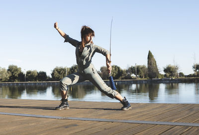 Confident female athlete with sword performing martial arts with dedication by lake on sunny day