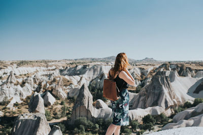 Rear view of young woman with backpack standing on landscape against clear blue sky during sunny day