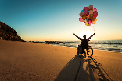 Silhouette mid adult woman with arms raised holding colorful balloons while sitting on wheelchair at beach during sunset