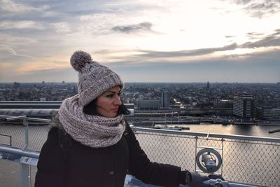 Woman looking away while standing by railing in city against sky during winter