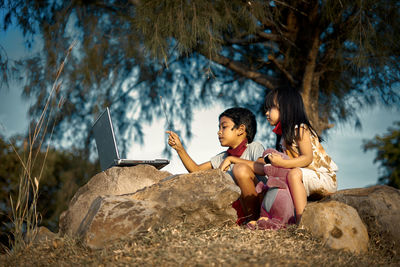 Siblings using laptop on rocks during sunny day
