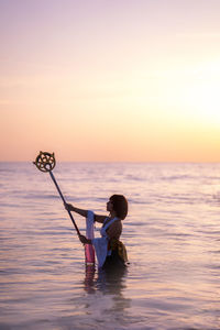 Man sitting in sea against sky during sunset