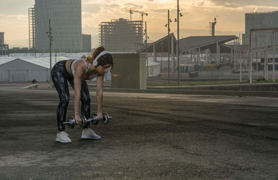 Woman listening music while lifting dumbbells in city during sunset