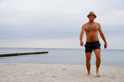 Shirtless young man walking at beach against sky