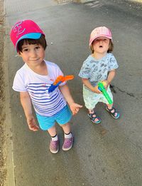 Two cute siblings standing on footpath wearing funny colourful hats and water guns